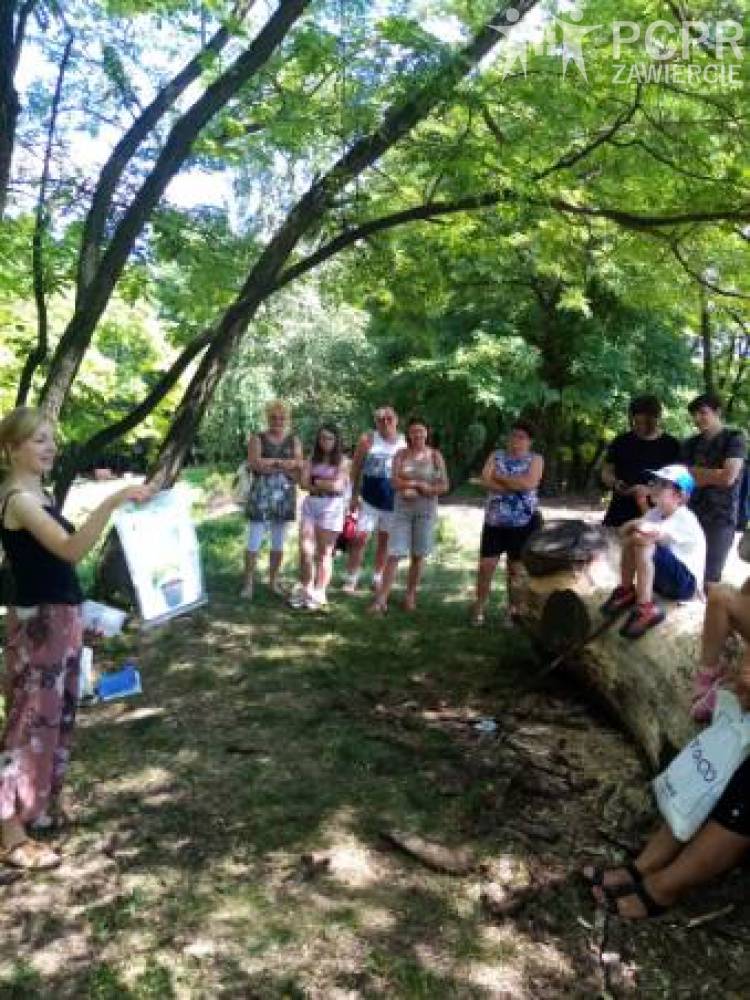 Zdjęcie: Liczna grupa osób stoi w parku przy drzewie i słucha wypowiadającej się kobiety